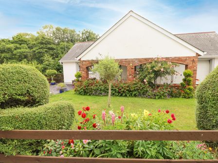 A garden with flowering plants and trimmed bushes in front of a white house with a brick archway at Charlecote House in Saundersfoot