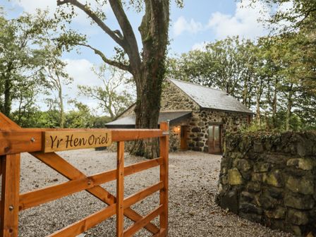 A wooden gate with Yr Hen Oriel sign leading to a stone house surrounded by trees at Yr Hen Oriel in Llanrug