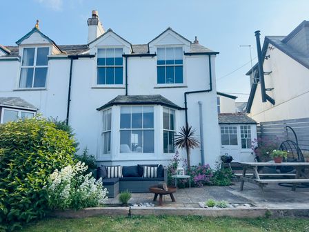 A white two-story house with large windows and a garden with outdoor seating and plants at 1 Tintagel in Port Isaac