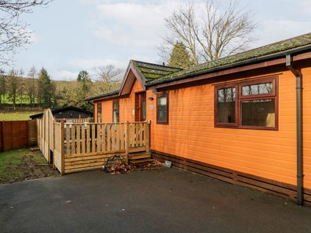 An orange wooden cabin with a fenced porch and trees in the background at Wansfell View in Limefitt Park near Troutbeck