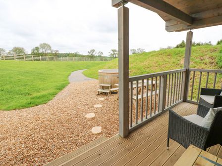 A wooden deck with two chairs and a railing overlooking a gravel pathway leading to a wooden hot tub and grassy area at Foxes Den in Winston near Staindrop