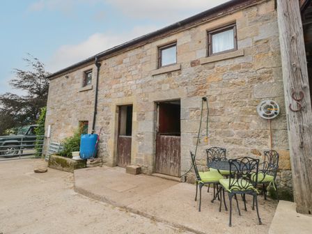 An outdoor patio area with iron table and chairs with green cushions beside a stone building with wooden doors and windows at The Granary in Rothbury