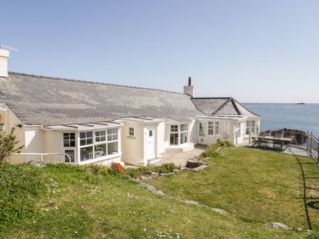 A white house with multiple windows and a gray roof overlooking the sea with a grassy yard and outdoor furniture at Craig Y Wylan in Bull Bay