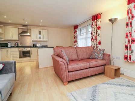 A living room and kitchen with a red sofa gray armchair wooden floor and floral curtains at 1 Tregonwell Court Minehead