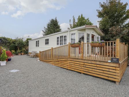 A mobile home with a wooden deck and flower baskets on gravel at Tegfryn Bach in Llangoed near Beaumaris