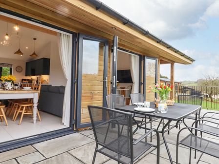 A patio with a black metal table and chairs set with flowers and dishes next to an open living dining area at Woodpecker Lodge in Butterleigh