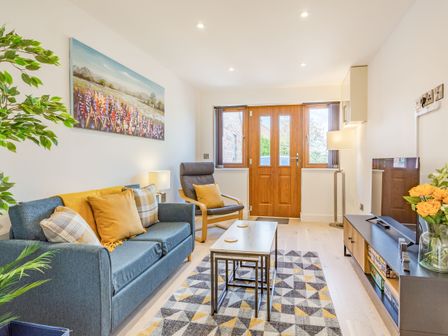 A living room with a blue sofa yellow pillows a wooden armchair a coffee table a patterned rug a TV stand with a television and a wooden door at The Belfry in Evesham