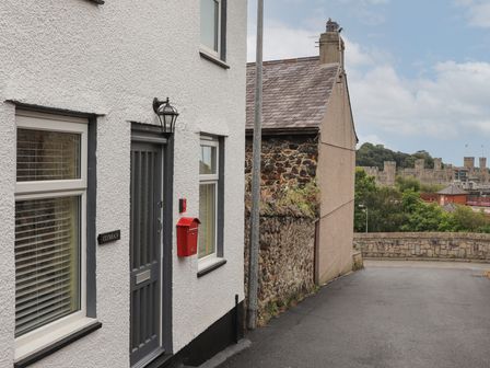 A street view with a white building with a grey door and red mailbox next to a stone wall and a castle in the background at Clydfan in Caernarfon