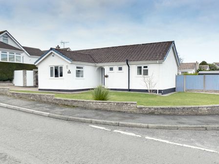 A detached single-story house with white walls and brown roof tiles on a corner lot with grass and a low brick wall in front at Gwersyllt in Benllech