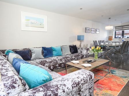A living room with a sectional sofa with blue and gray pillows a coffee table with flowers and dishes and a dining area with a glass table and gray chairs at Harbour Cottage in Dartmouth