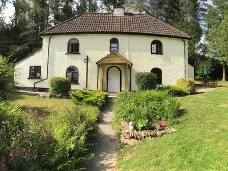 A house with a tiled roof and arched windows surrounded by garden plants and trees at Barn Owl Cottage in Wheddon Cross