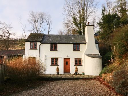 A white cottage with a stone roof and wooden door surrounded by bushes and trees at Hedgehog Cottage in Wheddon Cross