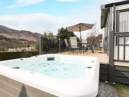 An outdoor hot tub next to a deck with a table umbrella and chairs with mountains in the background at Red Kite Lodge in St Fillans