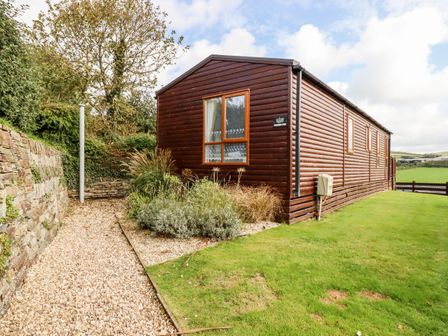A brown wooden house with a window surrounded by grass and gravel path by a stone wall at Orchard View in Bossiney near Tintagel