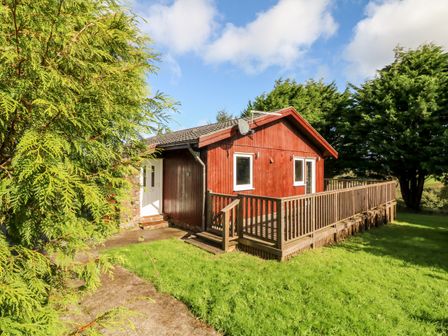 A small red wooden house with a deck surrounded by green grass and trees at Stonewood Lodge in Woolsery