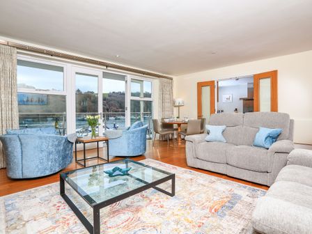 A living room with blue chairs a glass coffee table and a dining table near large windows overlooking water at 15 Dart Marina in Dartmouth