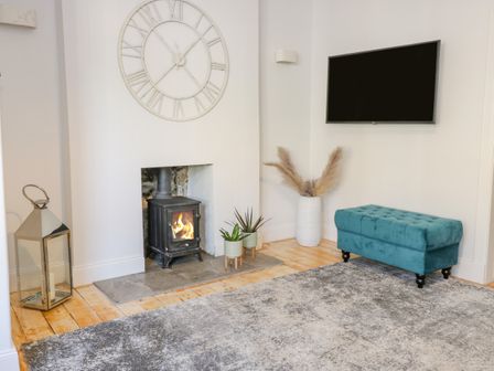A room with a small fireplace with fire a large wall clock a teal bench a wall-mounted television and plants at The Lazy Seal in Spittal