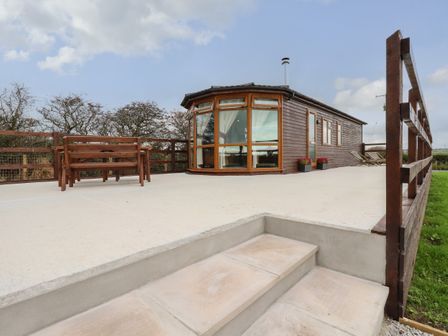 A wooden cabin with large windows on a concrete patio with wooden railing and outdoor bench seating at Sycamore Lodge Porkellis near Helston