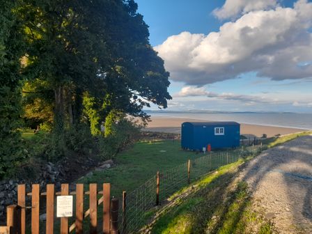 A blue shepherds hut near a sandy beach with trees and a wooden fence at Seashore Shepherds Hut @ Moat Farm in Aldingham near Ulverston