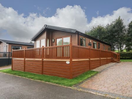 A brown wooden cabin with a surrounding deck and railing near a paved driveway and grass at The Warren in Hellifield