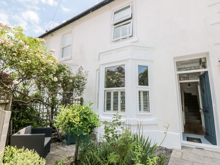 A front garden with outdoor seating and plants in front of a white house with a blue door at Bay Tree Cottage Dartmouth