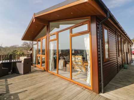 A wooden cabin with large glass windows and a deck with outdoor furniture at Meadow Lodge in Newton Ferrers