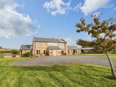 A stone farmhouse with multiple windows and a gravel driveway surrounded by green grass and a tree at Fontburn Farmhouse in Ewesley near Rothbury