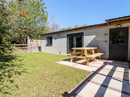 An outdoor area with grass a wooden picnic table and a building with a door and windows at Stall 2 - The Stables Brighton near Summercourt