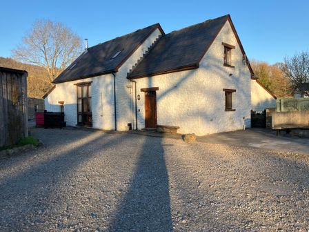 A white stone house with wooden doors and windows on a gravel driveway with trees in the background at Maes Y Berllan Barn in Gilwern