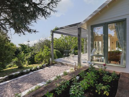 A garden with flowers next to a gravel path and a wooden porch with table and chairs at Meadow Lodge @ Seawardstone in Blackawton