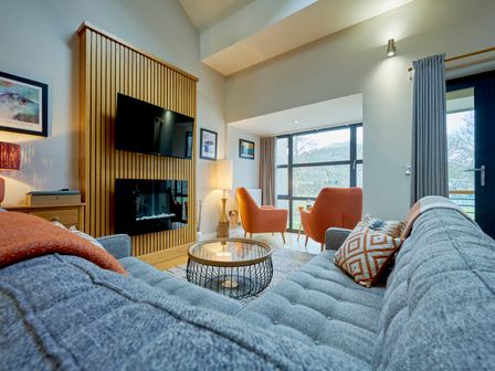 A living room with a grey couch orange chairs a round coffee table a mounted TV and a large window at Riverside Park 6 in Keswick