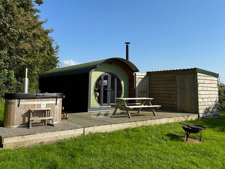 A wooden pod with curved roof next to a wooden hot tub and shed on a wooden deck with a picnic table and a fire pit on grass at Lime Kiln in Blagdon