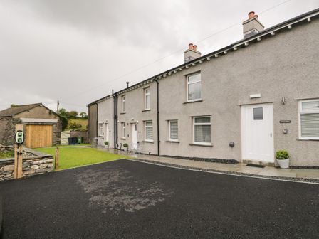 Exterior view of a row of gray houses with white doors and windows a small grass area a driveway and a stone building at Molly's Pantry Oxen Park near Ulverston