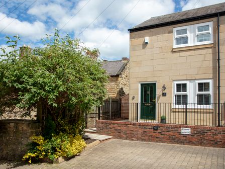 The exterior of a two storey stone house with a green door and white framed windows with a brick wall and a paved driveway and plants on the side at 1 Wesley Mews in Alnwick