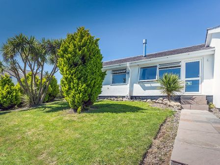 A grassy front yard with shrubs and a paved pathway leading to a white house with windows at 118 Cae Du in Abersoch