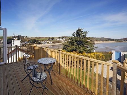A wooden balcony with a metal table and two chairs overlooking a beach and houses at Arwelfa Lodge in Abersoch