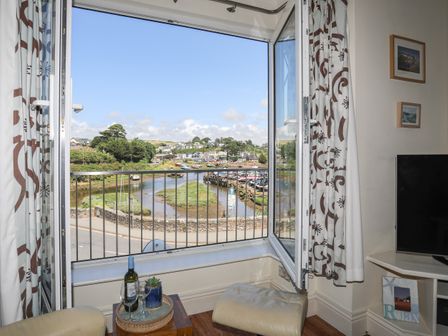 An open window with a view of a river harbor and houses in the distance inside a room with a small table and a chair at Harbour View in Abersoch