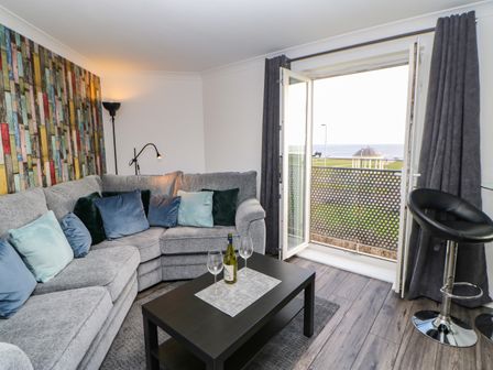 A living room with a grey sectional sofa and multiple cushions a black coffee table with a bottle and wine glasses an open door with dark curtains and a view of a gazebo and ocean at Sandy Corner in Blyth Northumberland