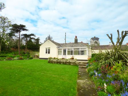 A white single story house with a large green lawn and flower beds in a garden at Hafod Cottage in Cemaes Bay