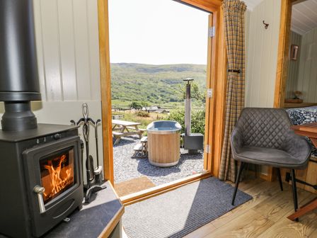 An interior view of a room with a wood burning stove a chair and an open door leading to an outdoor wooden hot tub and picnic table with hills in the background at Llethr - Shepherds Hut in Llanbedr
