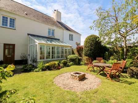 A garden with a grassy lawn a round stone fire pit wooden table and chairs and a conservatory at Home Farm Cottage in Fremington Devon