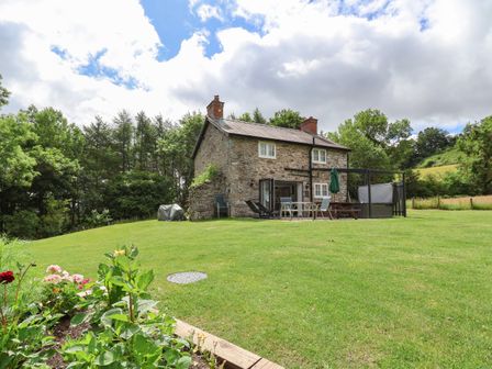 A stone cottage with a chimney and outdoor furniture on a grassy lawn surrounded by trees at Cae Iocyn in Chirk