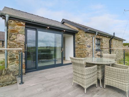 A stone house with sliding glass doors and outdoor wicker table and chairs on a wooden deck at Tyddyn Bach in Rhydwyn