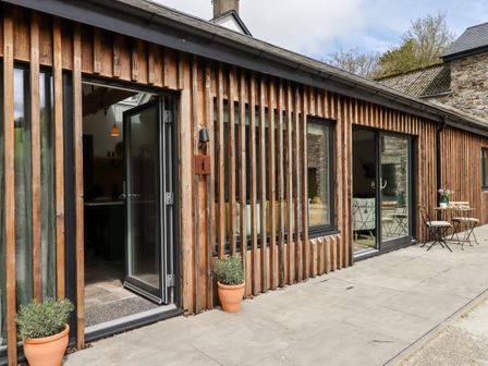 An exterior view of a wooden building with glass doors and windows two potted plants and an outdoor table with chairs at 1 Caddaford Barns in Buckfastleigh