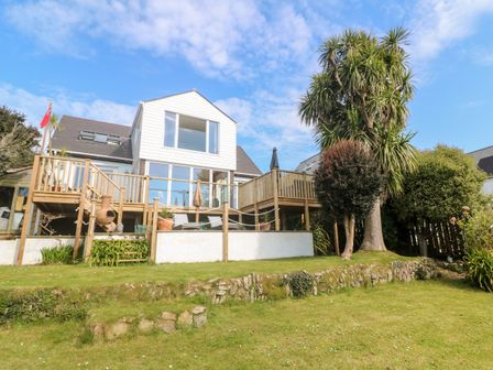 A white two-story house with a wooden deck and glass doors overlooking a garden with grass and trees at Trewartha in Praa Sands