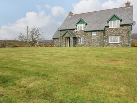 A stone house with a gabled roof and green trim on a grassy lawn with a tree and a swing set at Kiltymon Cottage Ahakista near Bantry County Cork