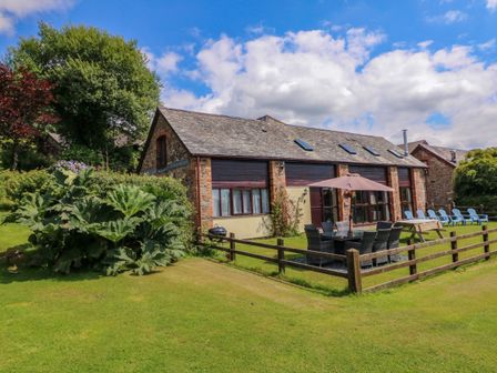 A stone barn with outdoor dining table umbrella and lawn chairs on a grass lawn at Apple Barn in South Molton