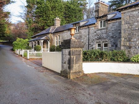 Stone house with white fence and gate along a road with trees and bushes at 1 The Stables in Grange-Over-Sands