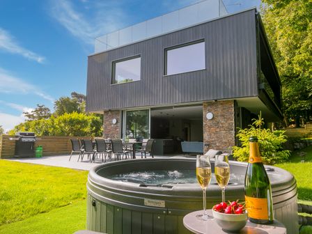 An outdoor patio with a dining table and chairs next to a modern house with large windows and a hot tub with champagne and cherries nearby at Sparrows Nest in St Ives