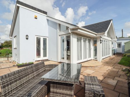 A patio area with a glass outdoor table and wicker furniture outside a white house with large glass doors at Avalon in Lamphey